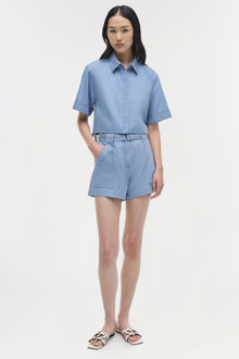 Light blue denim button-up shirt with short sleeves and cropped length, styled with matching denim shorts.