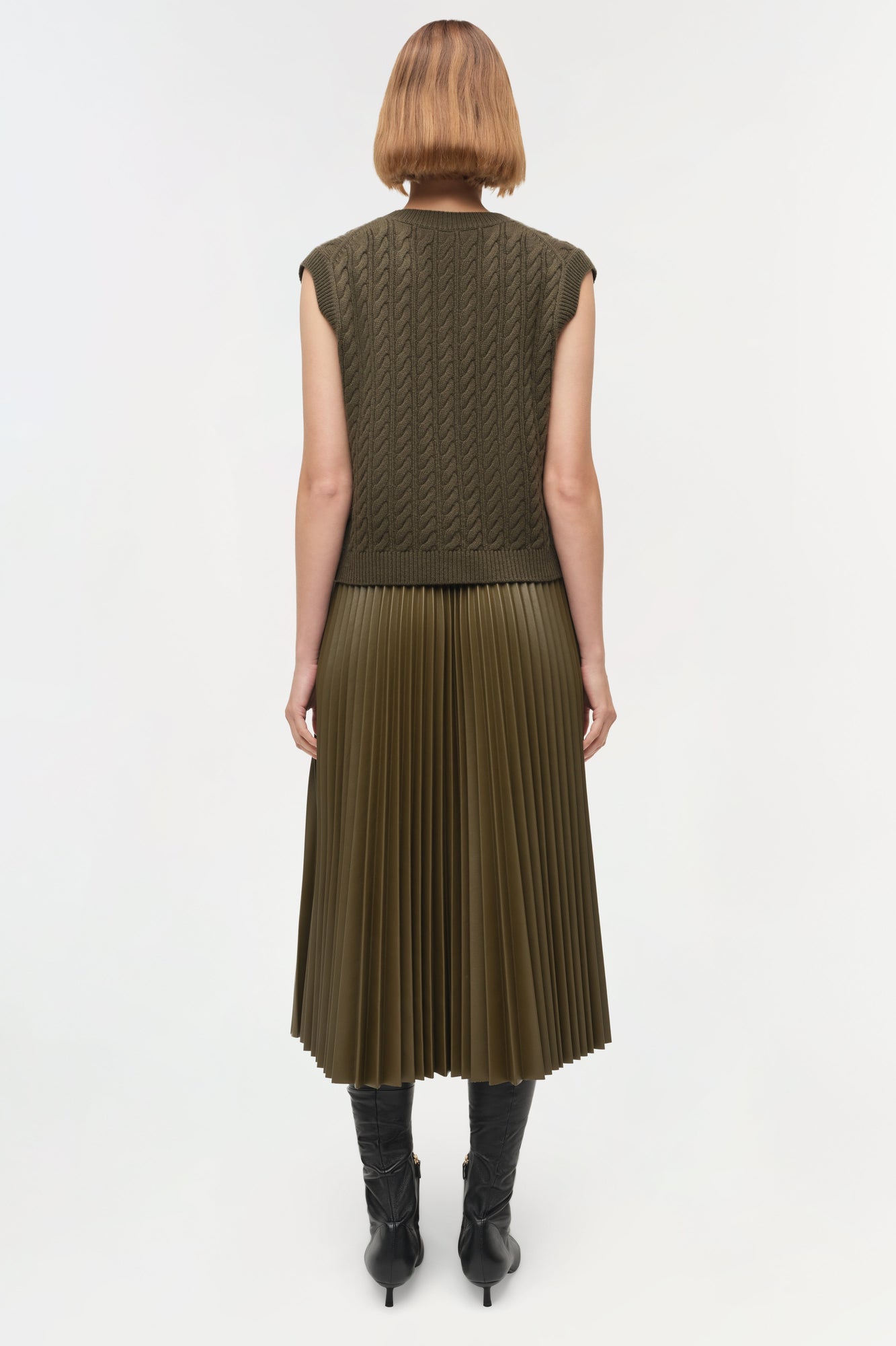 Olive green sleeveless cashmere vest with cable knit pattern, shown from back on model wearing pleated skirt.