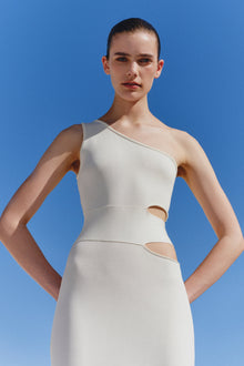 White one-shoulder dress with cutout details at the waist and side, worn by model against blue background.