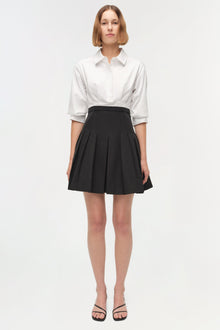 White collared shirt tucked into black pleated mini skirt with three-quarter sleeves.