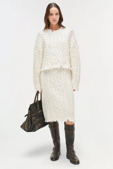 Cream cable-knit midi skirt with distressed hem edges, worn with matching sweater and black boots.