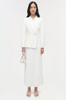 Woman wearing cream pleated blazer midi dress with long sleeves, paired with white sandals and matching handbag.