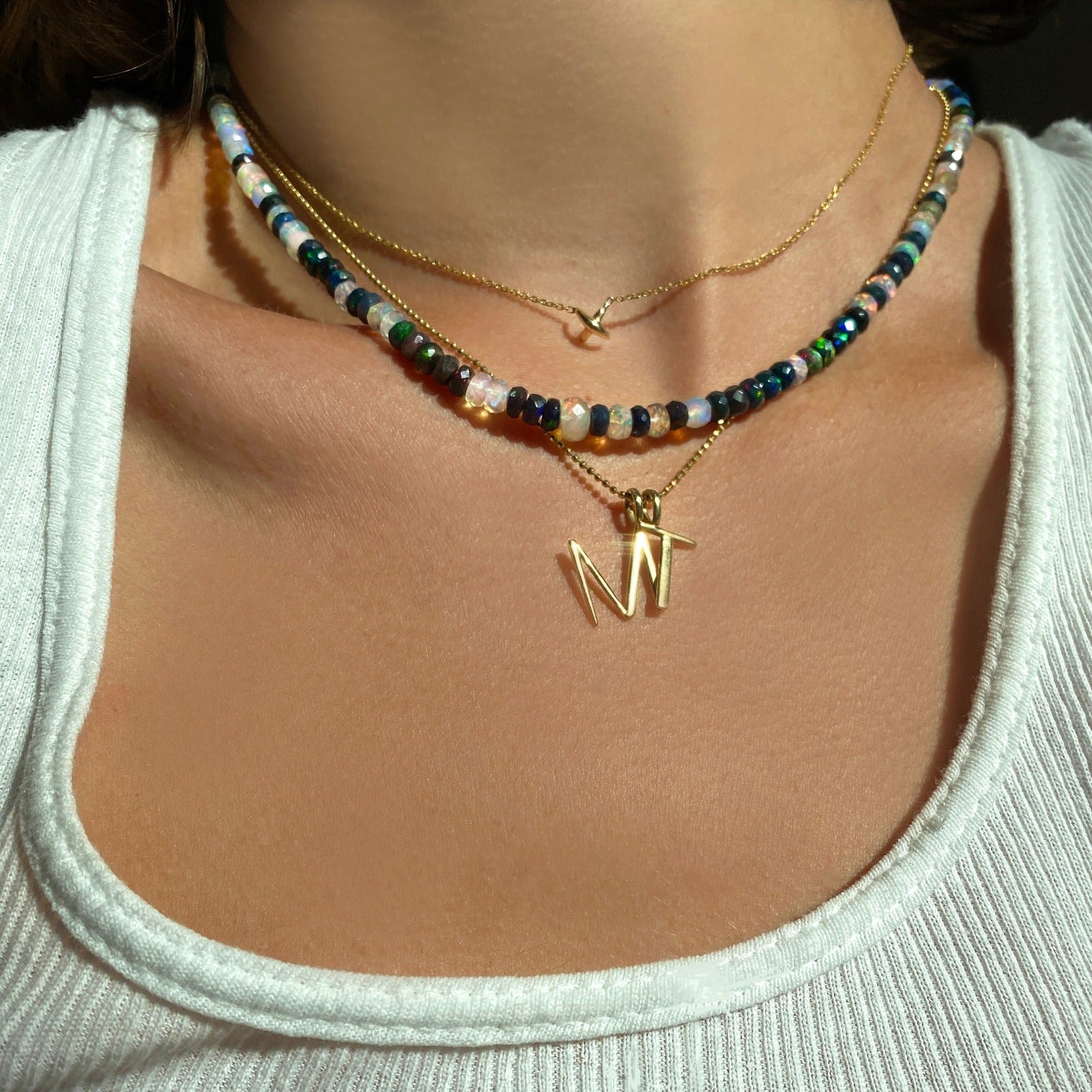 Shimmering beaded necklace made of faceted opals in shades of fiery pastels, grey, teal, and black on a gold linking ovals clasp. Styled on a neck layered with a stitch necklace, bead chain necklace, M and T letter charms.