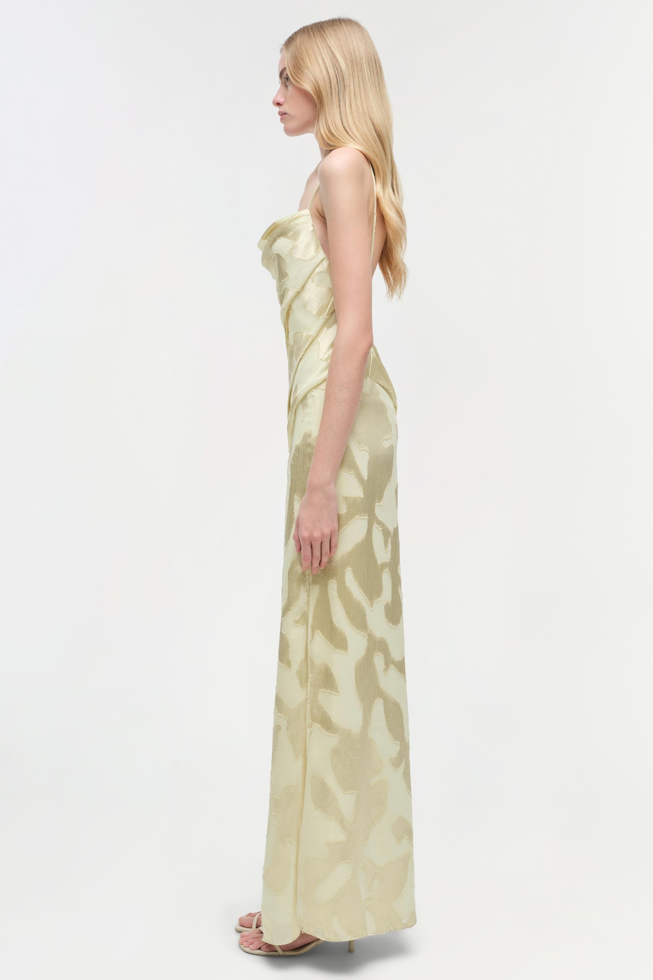 Pale yellow sleeveless floor-length gown with abstract leaf pattern shown on model from side profile.