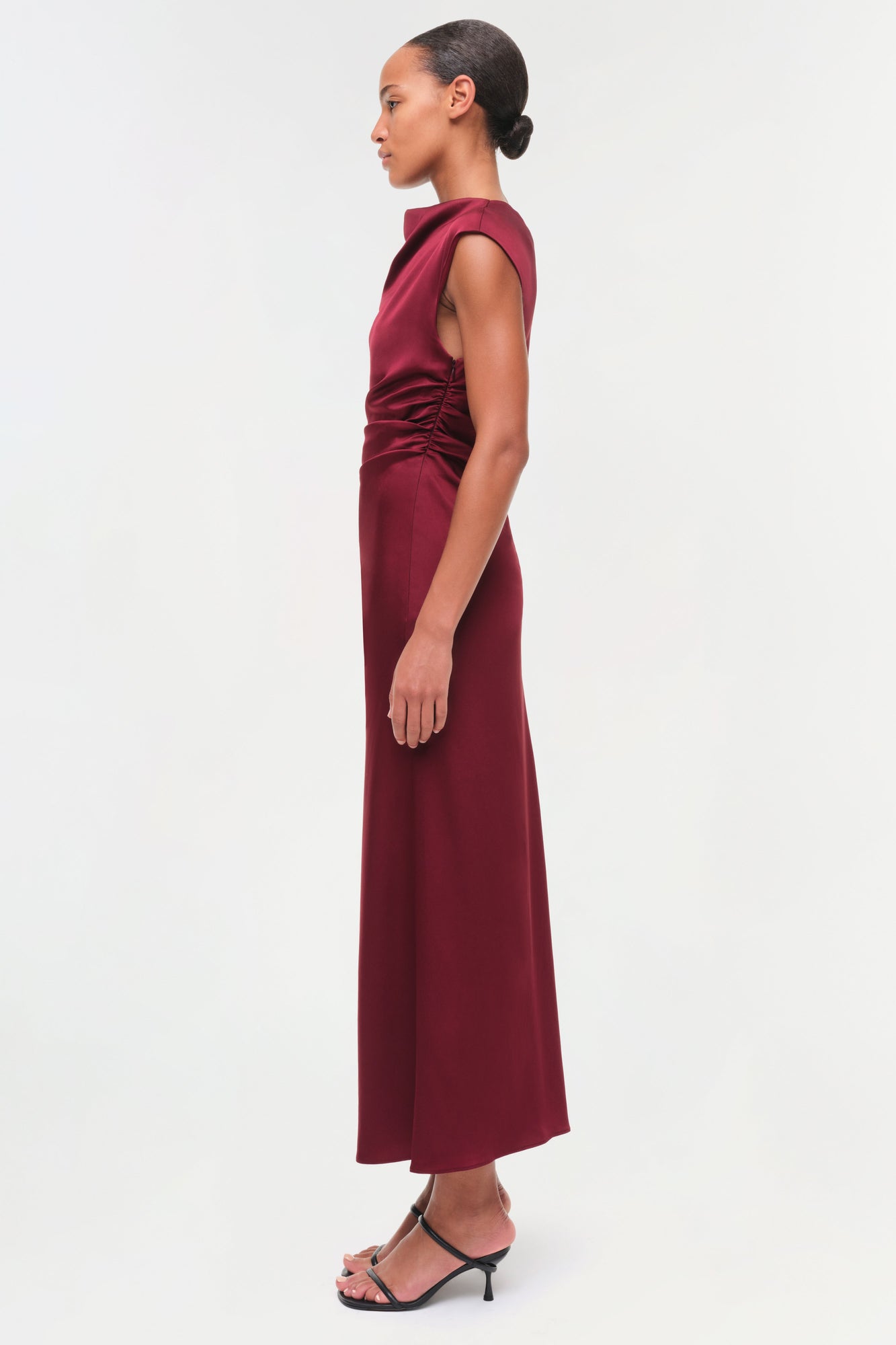 Burgundy sleeveless satin midi dress with ruched side detailing, shown on model from side profile.
