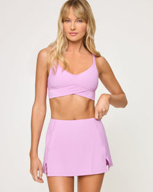 Close-up front view from the waist up of the LSPACE Unmatched Bra in mauve on a model against a white background, highlighting the crossover bra band and the cream crossover waistband of the matching mauve skort.