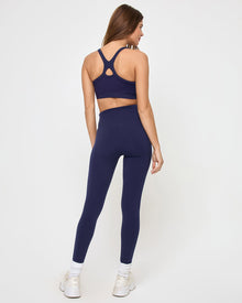 Back view of the LSPACE Uppercut Bra in midnight blue on a model against a white background, showing the crossback X-strap detail of the midnight blue sports bra, paired with matching midnight blue high-rise active pants, white sneakers and socks.