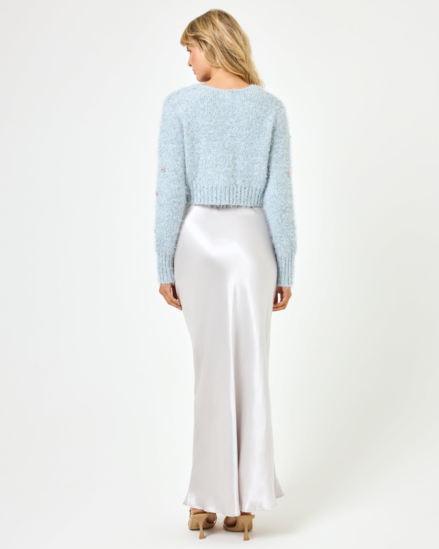 Back view of a model wearing the LSPACE Sweet As Honey Skirt in silver, showing the smooth satin back and A-line silhouette, styled with a pale blue fluffy cardigan and heeled sandals on a white background.