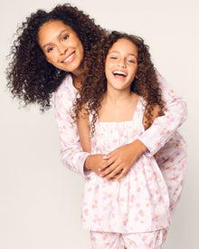 Two people with curly hair, wearing the luxurious Petite Plume Girls Twill Charlotte Short Set in Seashell Splendor, are smiling and embracing in front of a plain background.