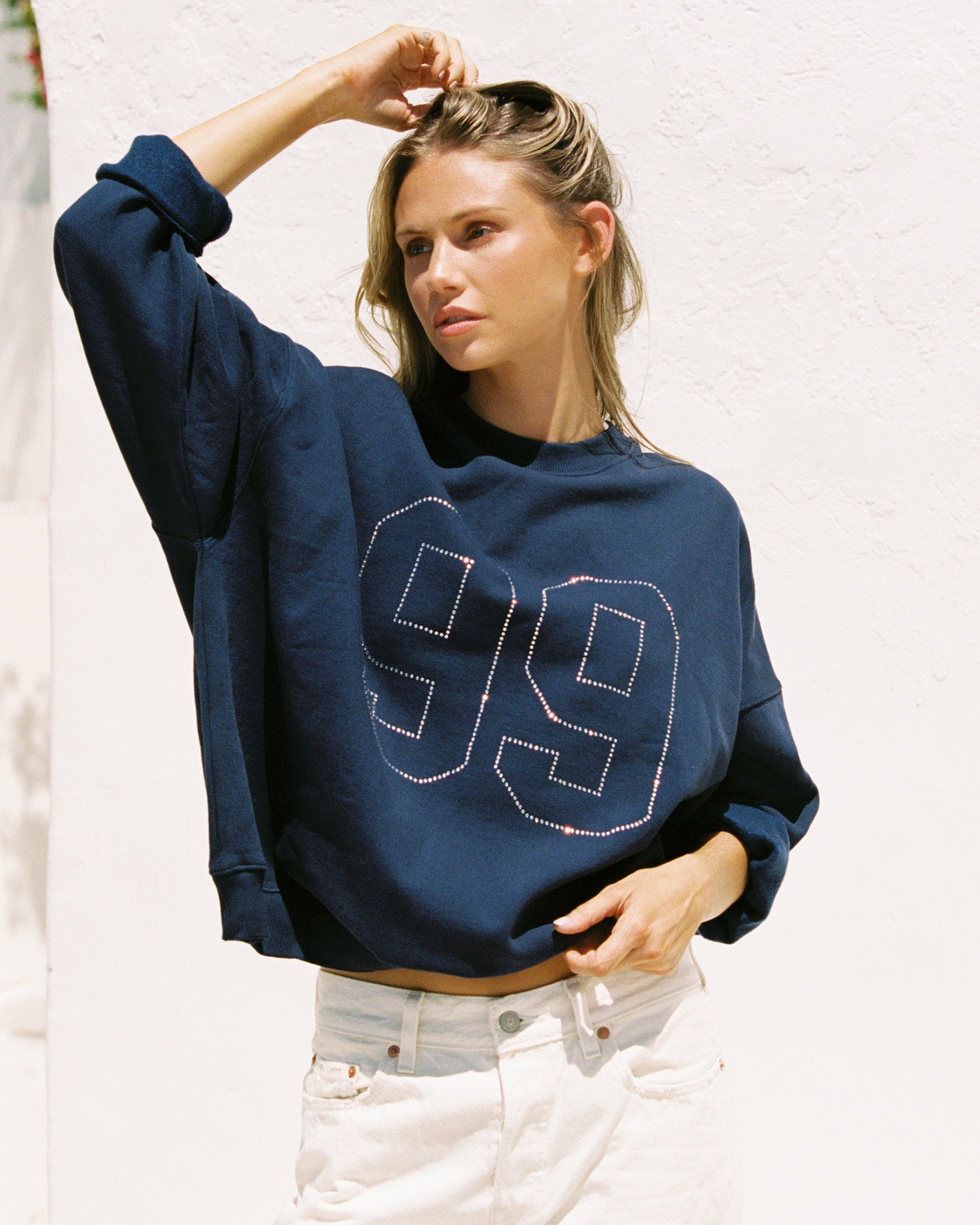 Lifestyle editorial shot of a model wearing the LSPACE Legend Sweatshirt in midnight blue, an oversized cropped sweatshirt with a rhinestone number graphic, paired with white denim jeans against an outdoor white wall.