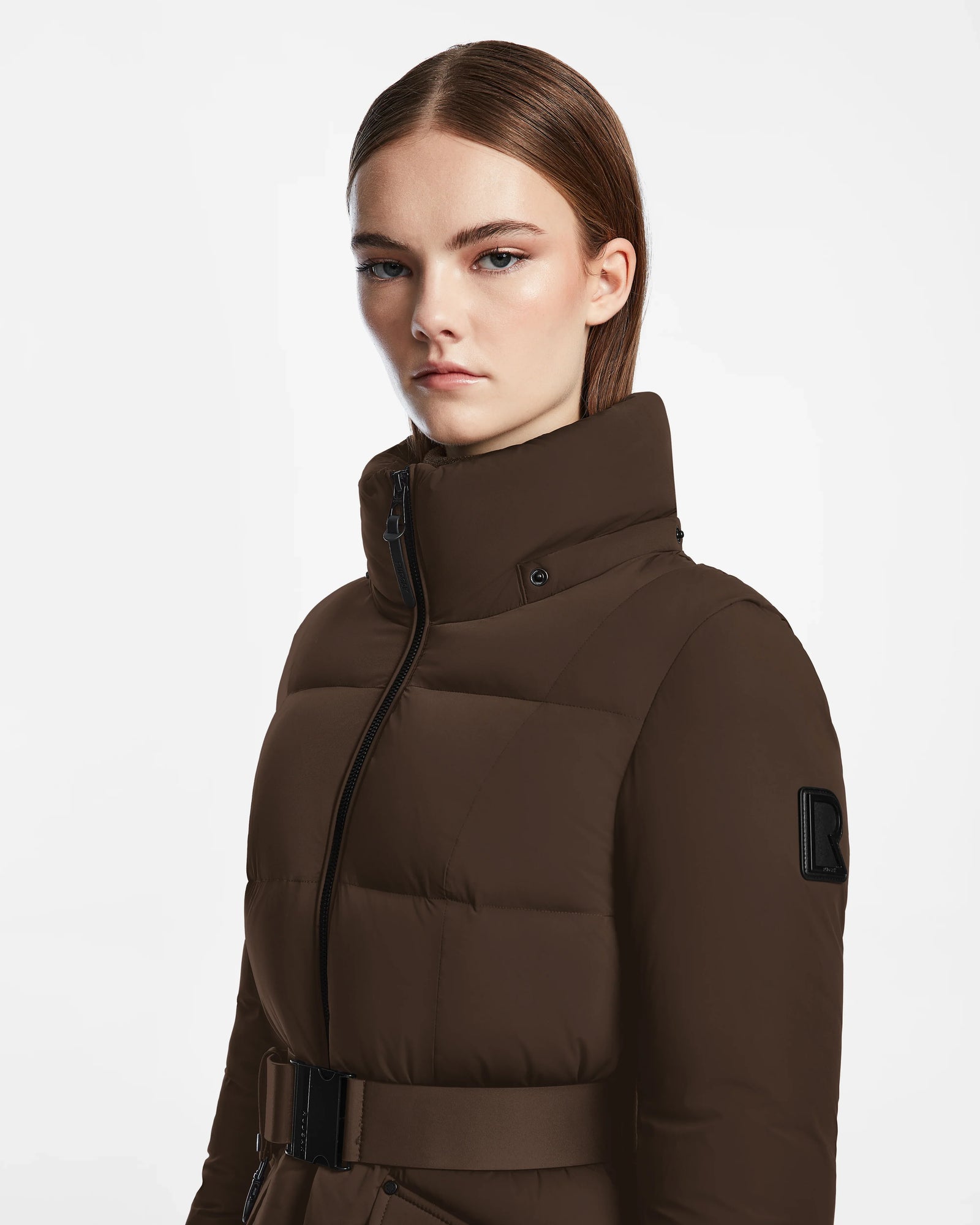 A woman with straight brown hair and light makeup wears the Rudsak ADELINE - 8124544 Mocha quilted short down puffer jacket with a belt, high collar, and black sleeve patch, standing against a plain white background.