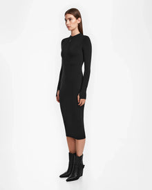 A woman stands sideways against a white background, wearing the AGNES - 8524551 Black bodycon stretch dress by Rudsak with black ankle boots. Her straight brown hair is down and she faces forward with a neutral expression.