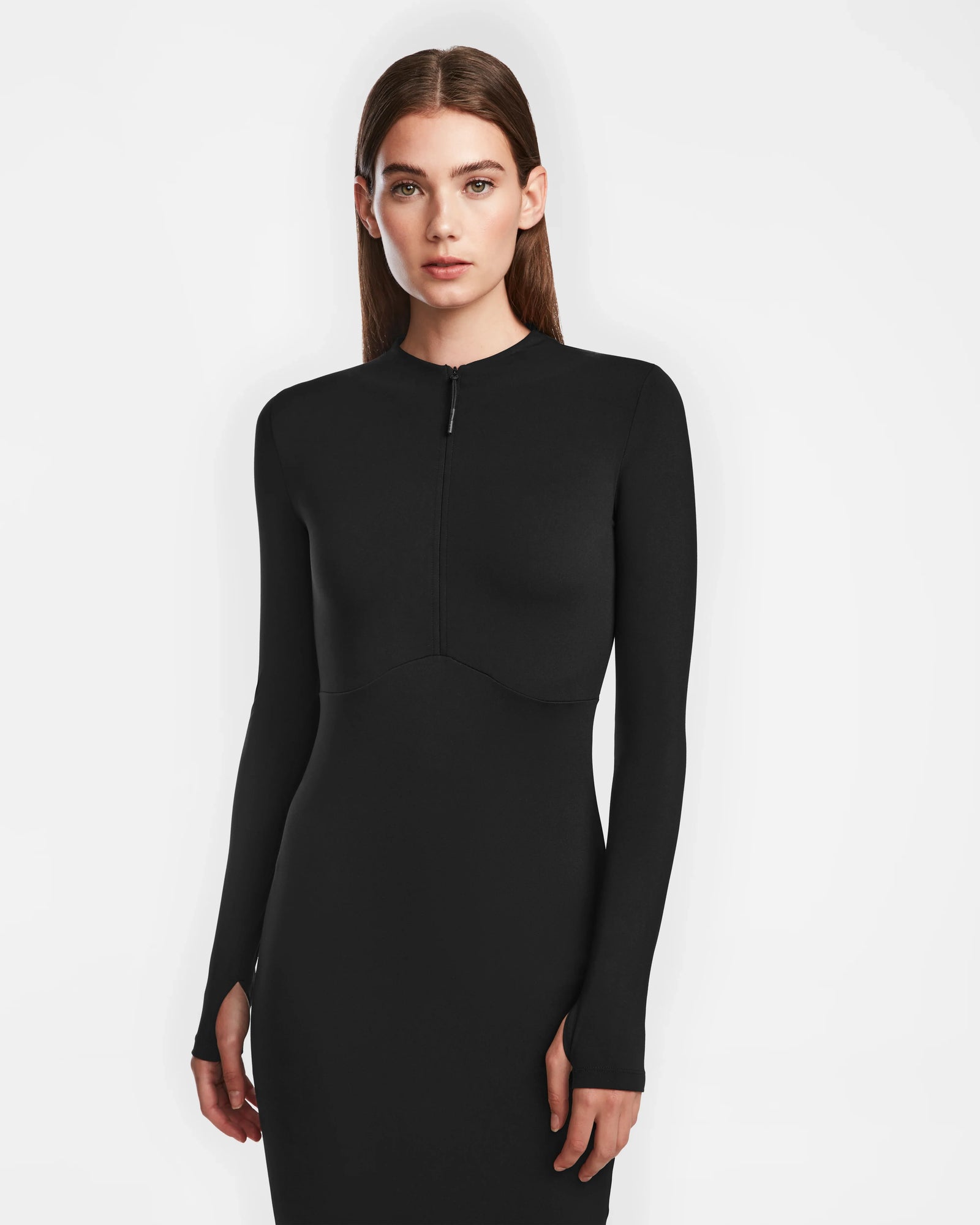 A woman with straight brown hair wears the Rudsak AGNES - 8524551 Black fitted womens bodycon dress with a short zip-up front, standing against a plain white background.