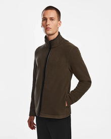 A man with short hair wears the Rudsak ARAGON - 6524539 Army mens fleece full-zip jacket with vegan leather trims and black pants, standing against a plain white background with his hands in his pockets, looking slightly to the side.