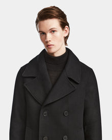 A young person with fair skin and light brown hair, wearing the Rudsak ARTHUR - 6124521 Black premium wool blend double-breasted peacoat over a black turtleneck, stands against a plain white background, facing slightly right.