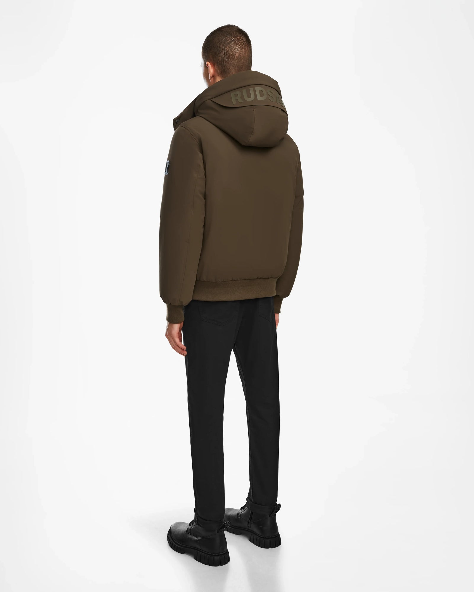 Facing away, a person wears the Rudsak ASH - 6124599 Army mens down bomber jacket with RUDSAK on the hood, paired with black pants and shoes—showcasing modern urban style against a plain white background.