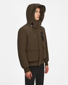 Wearing the Rudsak ASH - 6124599 Army men’s down bomber jacket with a brown hood, front and zippered chest pocket, and black pants, a person stands against a plain white background, highlighting modern urban style.