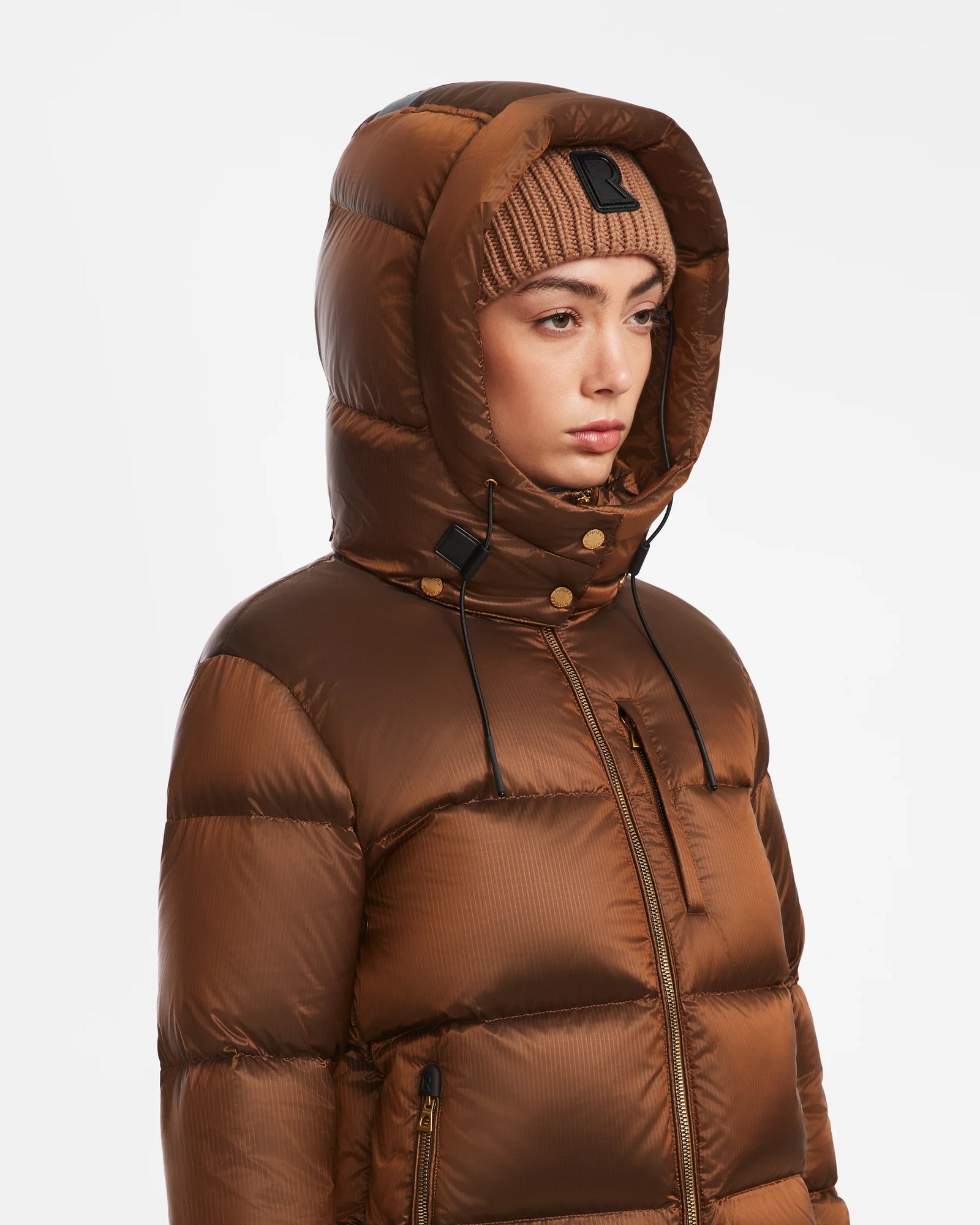 A person wearing the Rudsak ASHLYN S - 8124565 Antique Bronze womens down puffer jacket with the hood up and a beige knit beanie, looking to the side against a plain white background.
