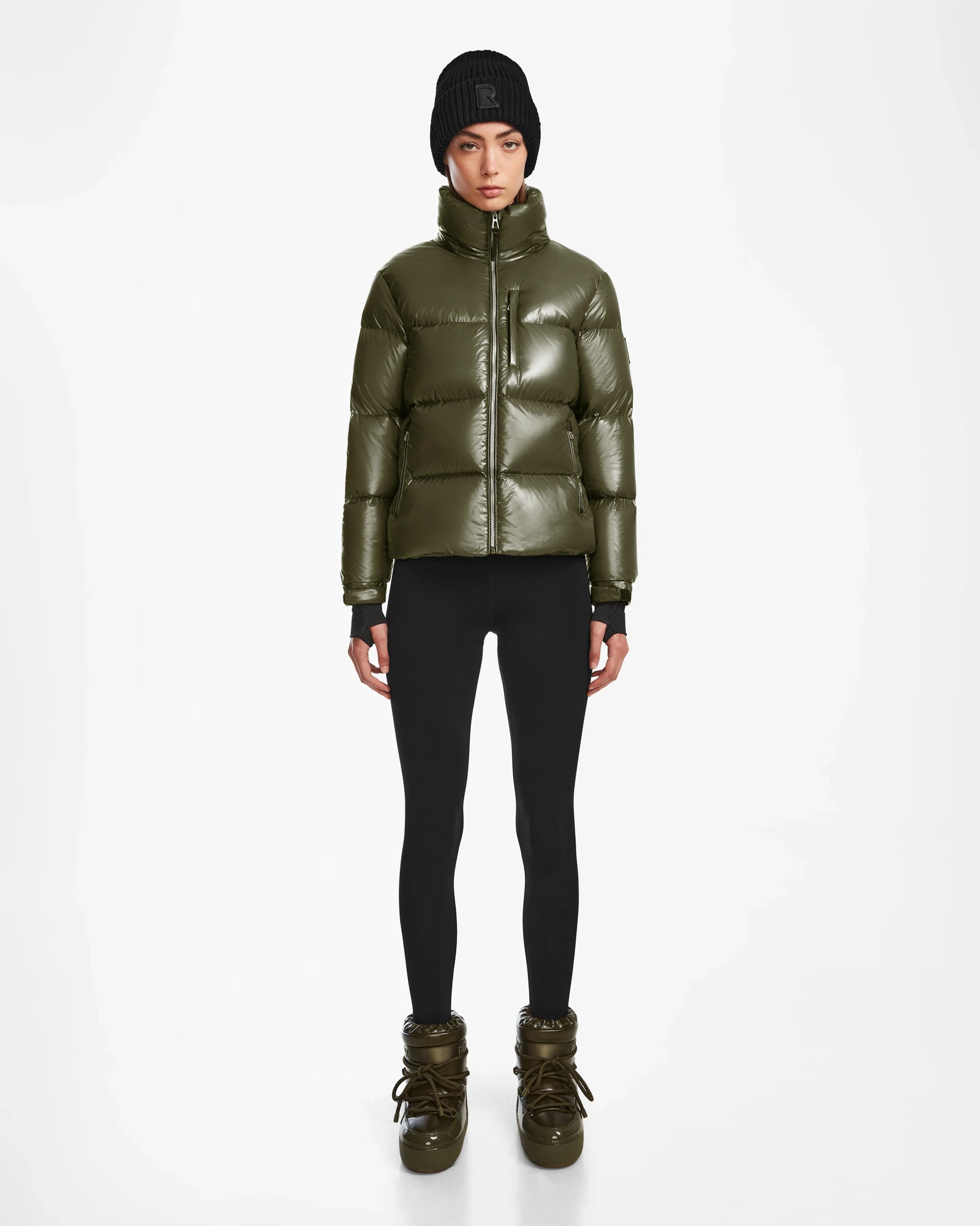 A person wearing the Rudsak ASHLYN S - 8124565 Loden Green puffer jacket stands against a plain white background, styled with black leggings, olive green winter boots, and a black knit beanie.