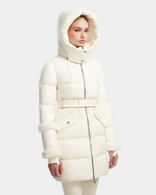 A woman wears the Rudsak AUDREY L - 8124585 Iceland knee-length down puffer jacket, made from recycled Stormshell fabric and RDS-certified down, with a belted waist, hood, and fur trim. She stands against a plain light gray background.