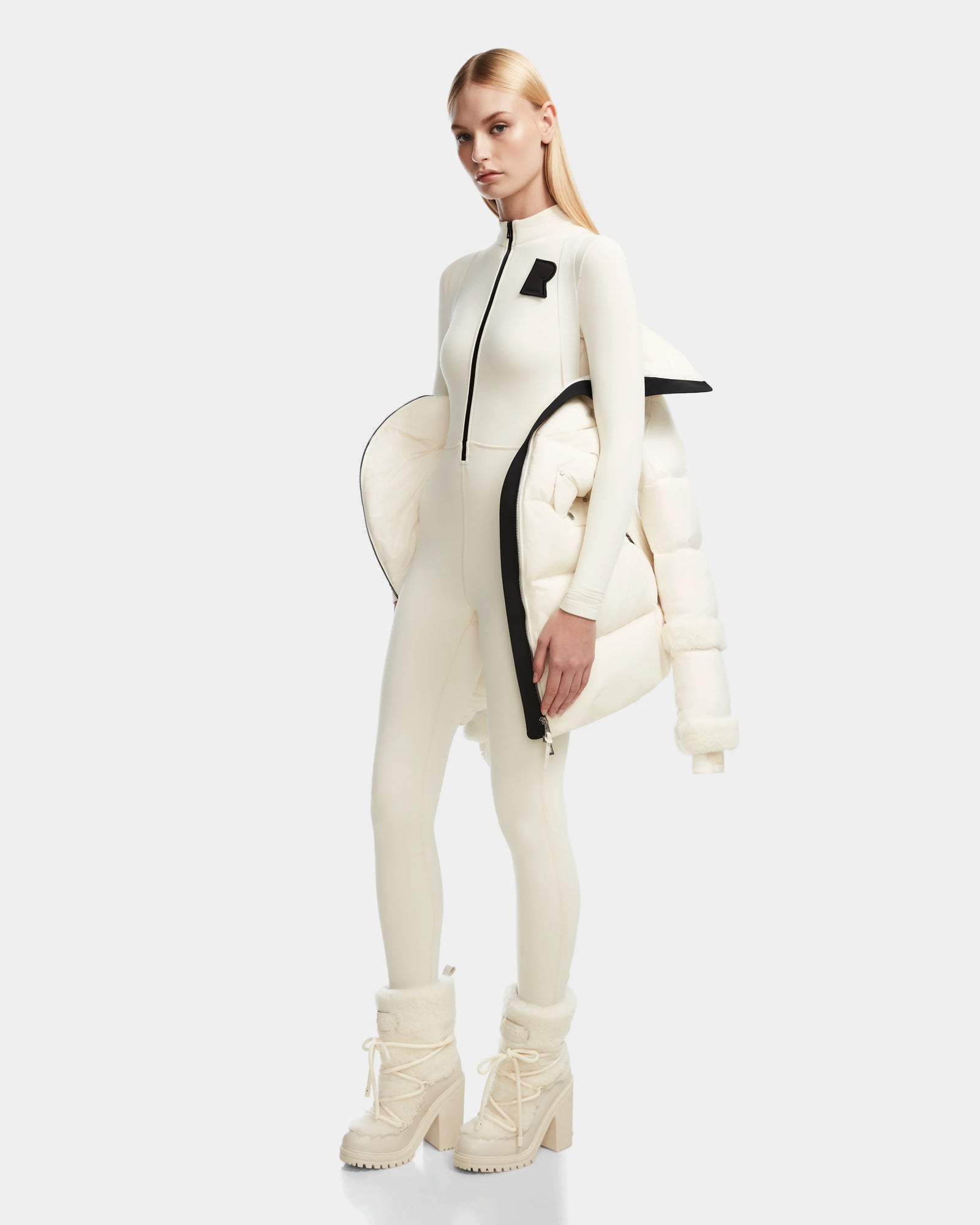 A woman poses against a plain backdrop in Rudsaks AUDREY L - 8124585 Iceland—a fitted ivory jumpsuit, matching high-heeled winter boots, and a knee-length RDS certified down puffer jacket draped over one shoulder. Her hair is sleek and straight.