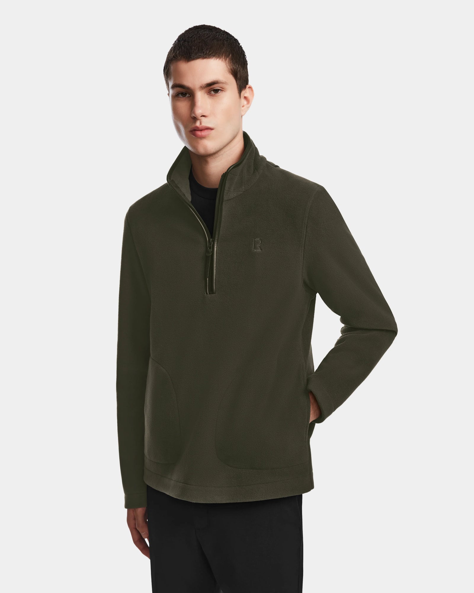 A young man wearing the Rudsak BECK - 6524538 Army men’s fleece pullover with vegan leather trims stands with hands in pockets against a plain white background.