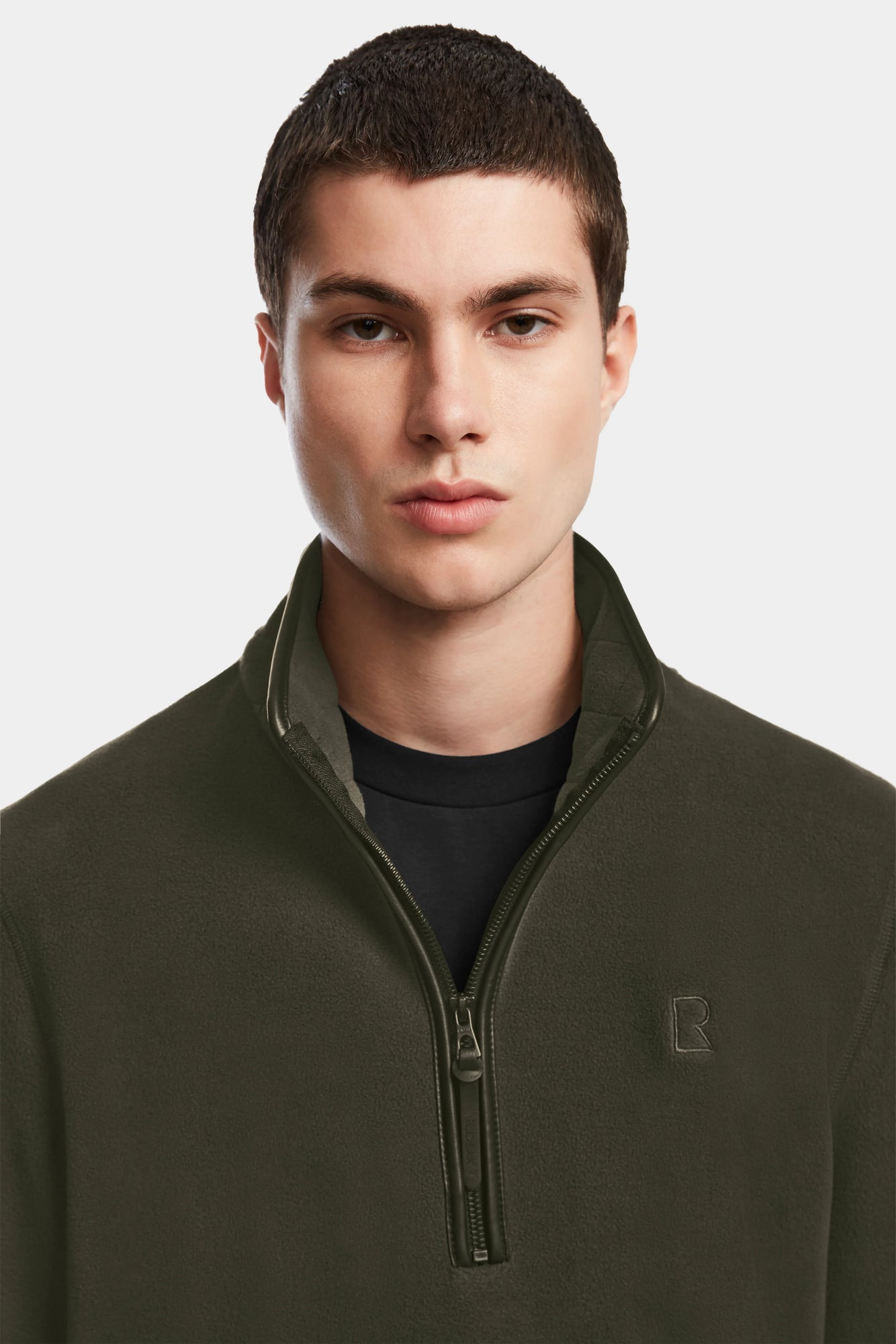 A young man with short dark hair wears the Rudsak BECK - 6524538 Army olive fleece pullover with vegan leather trims over a black shirt, standing against a plain light gray background.