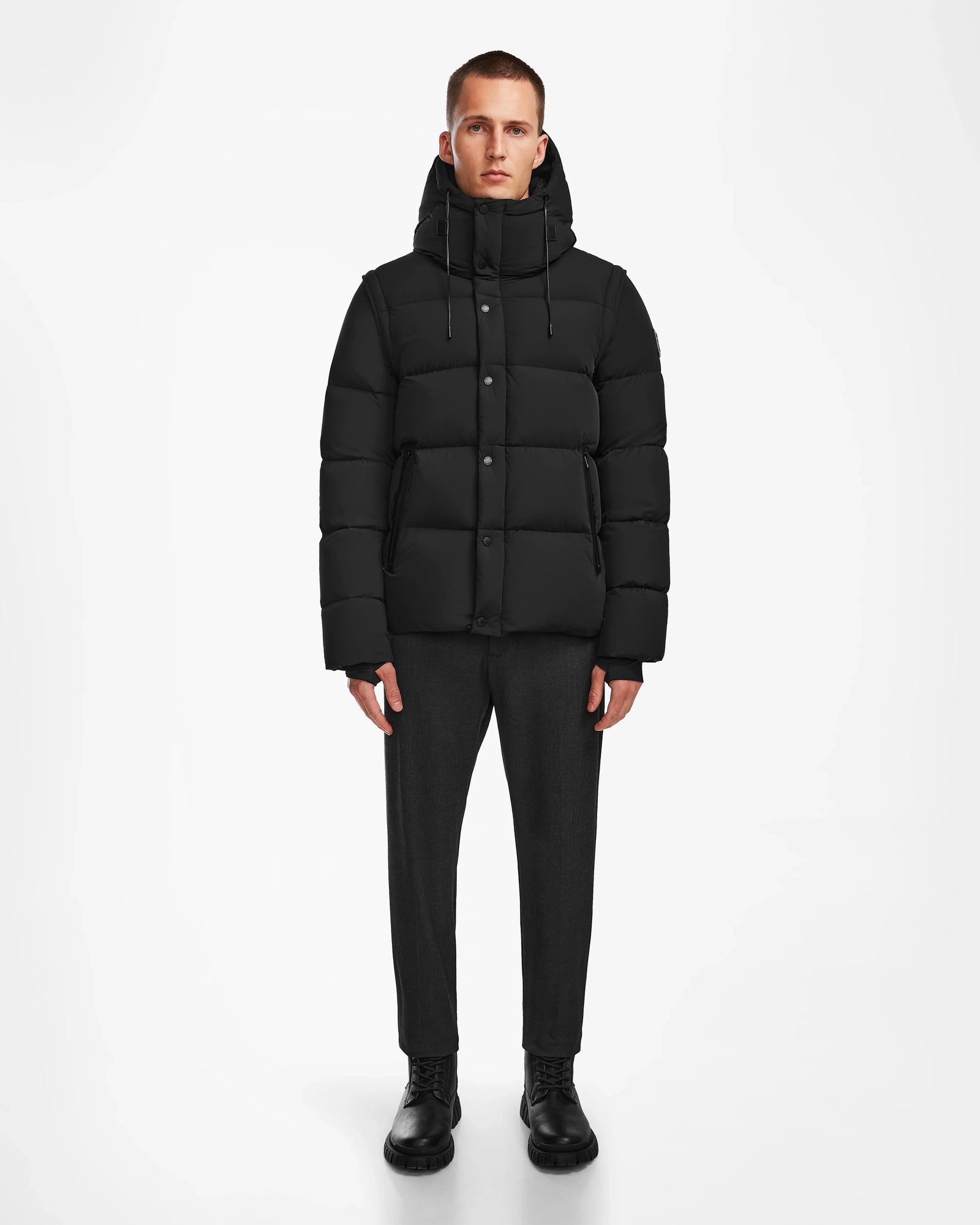 A man faces forward against a plain white background, wearing the Rudsak BOBBY - 6124506 Black mens down puffer jacket with a hood, paired with dark trousers and black shoes.