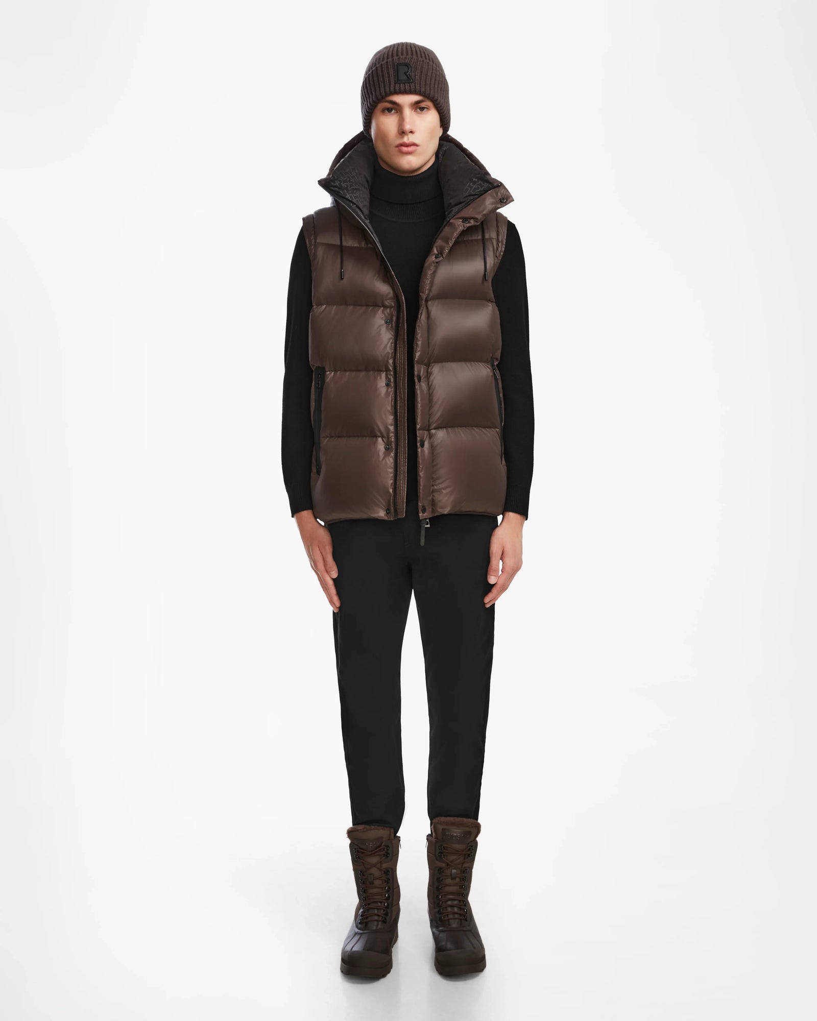 A person faces forward wearing the Rudsak BOBBY - 6124506 Espresso mens down puffer vest over a black turtleneck, black pants, brown boots, and a brown knit beanie against a plain white background.