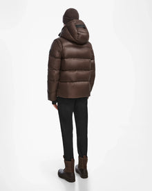 A person wearing the Rudsak BOBBY - 6124506 Espresso men’s quilted down puffer jacket, a brown knit hat, black pants, and brown boots stands facing away against a plain white background.