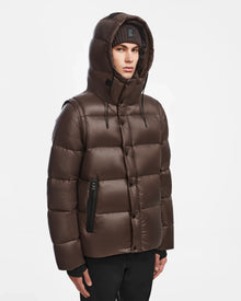 A person wearing the Rudsak BOBBY - 6124506 Espresso mens quilted down puffer with a hood and a black beanie stands against a plain white background, looking slightly to the side with a neutral expression.