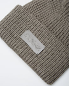 Close-up of the COLIN - 9424518 Cement beanie by Rudsak, a taupe unisex ribbed knit style featuring a rectangular patch embossed with the Rudsak brand name.