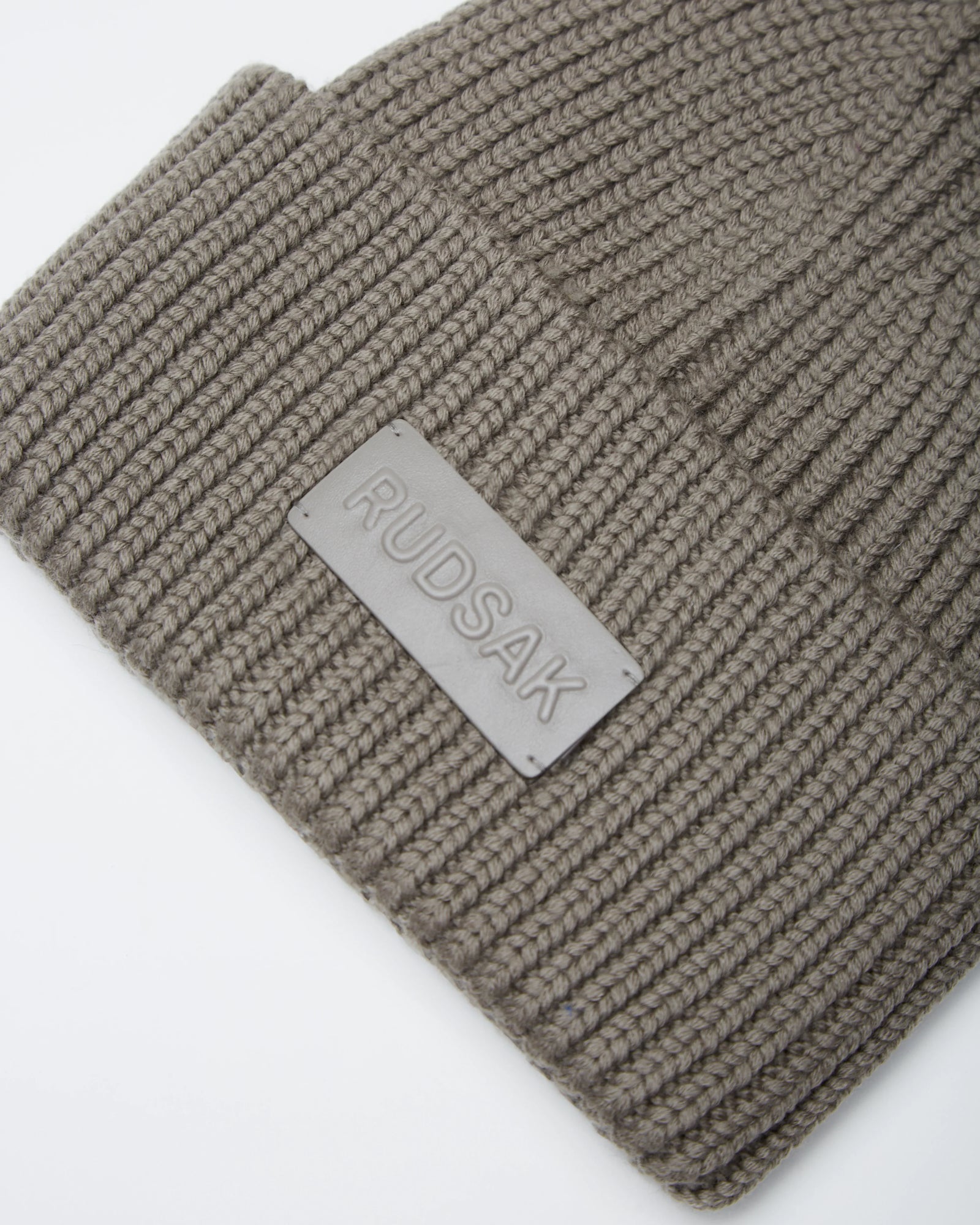 Close-up of the COLIN - 9424518 Cement beanie by Rudsak, a taupe unisex ribbed knit style featuring a rectangular patch embossed with the Rudsak brand name.