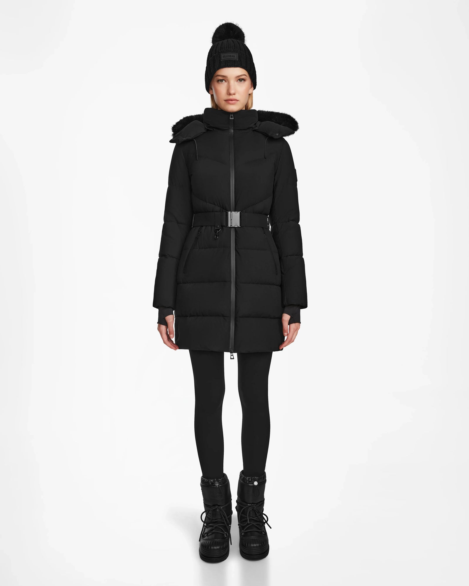 A person stands facing forward wearing the Rudsak CHRISTEL M-M - L124505 Black quilted winter coat with a belt, paired with black leggings, boots, and a pom-pom knit hat, against a plain white background.