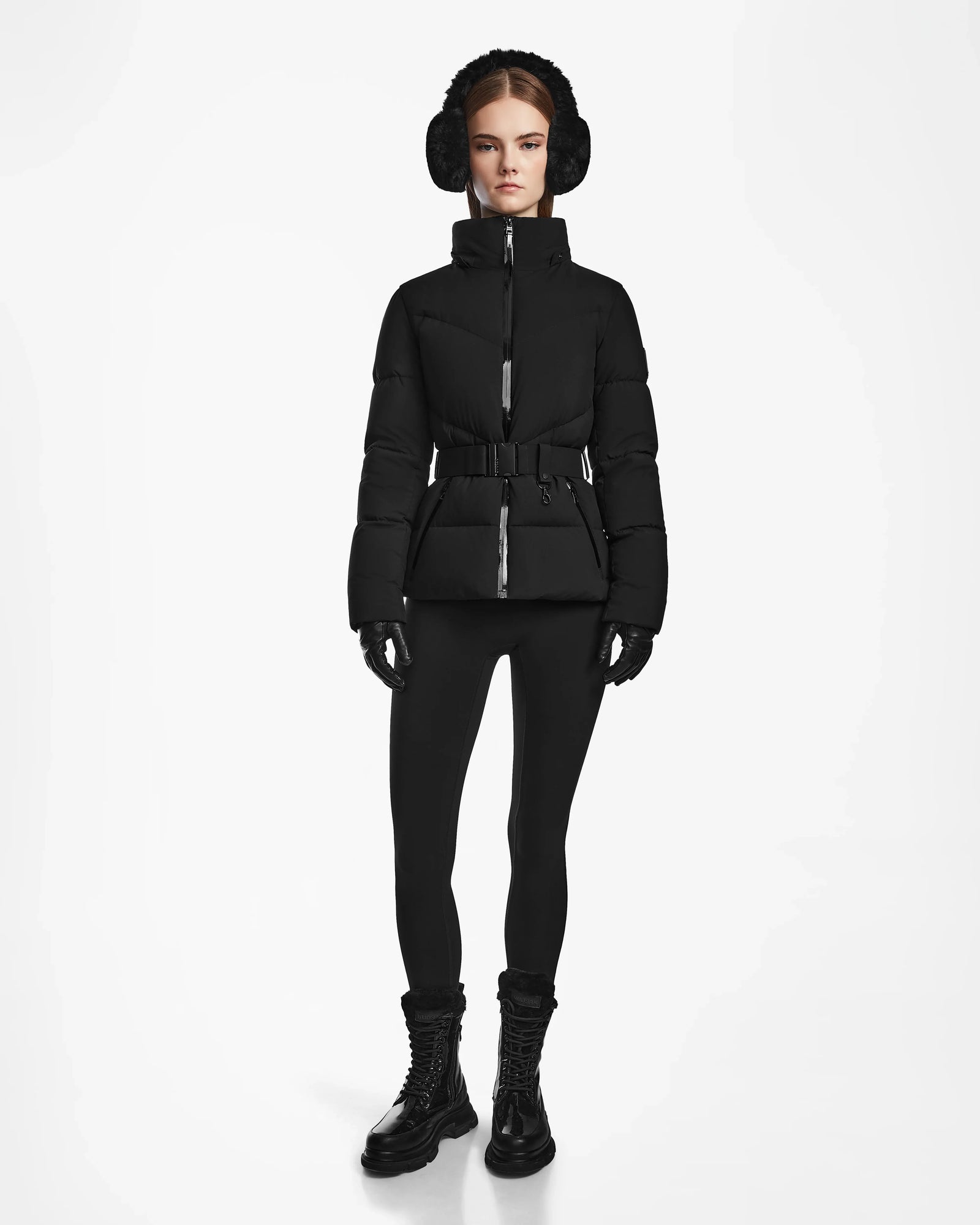 A woman stands facing forward, wearing the Rudsak CHRISTELLE M - L123525 Black women's quilted down puffer jacket with a fitted silhouette, styled with black leggings, earmuffs, gloves, and lace-up boots against a plain white background.