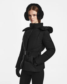 A woman wearing the Rudsak CHRISTELLE M - L123525 Black jacket with a belt, along with black earmuffs, gloves, and pants, stands against a plain white background looking straight ahead with a neutral expression.