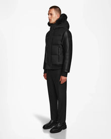 A man stands against a white background wearing the Rudsak DALLAS R - M123501 BLACK quilted down bomber with recycled leather sleeves, paired with black pants and shoes. He faces slightly to the side with his hands by his sides.