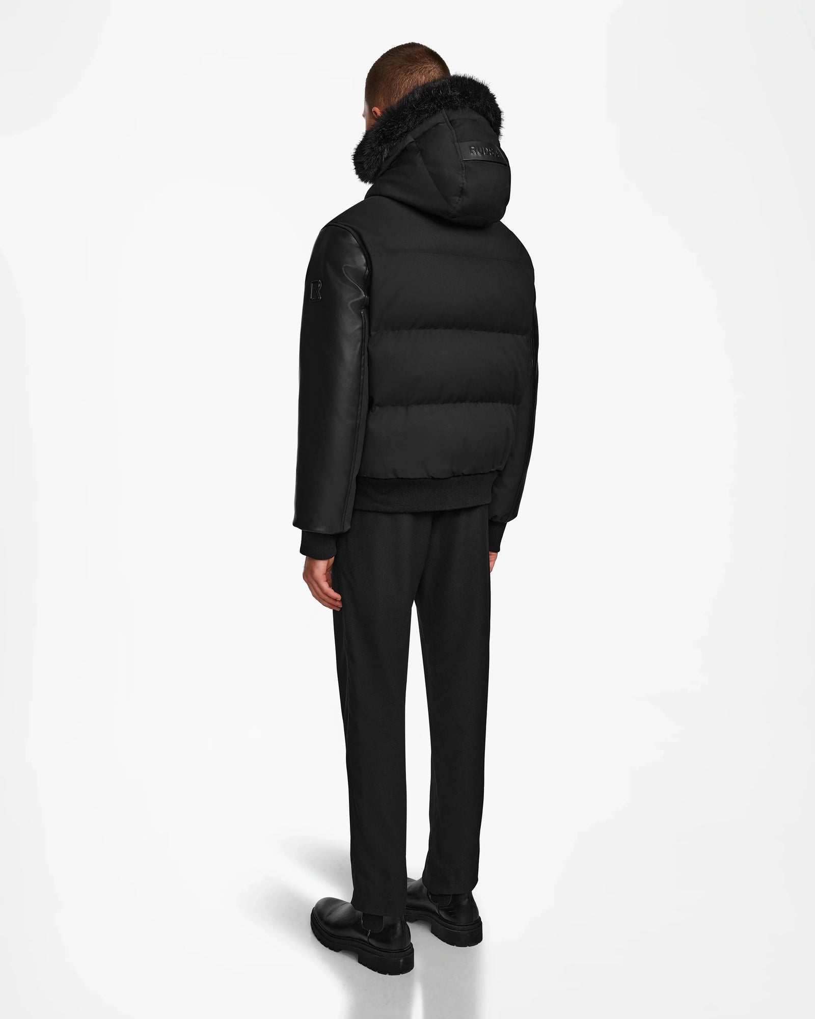 A person faces away, wearing the Rudsak DALLAS R - M123501 BLACK men’s quilted down bomber with recycled leather sleeves and a fur-trimmed hood, paired with black pants and shoes against a plain white background.