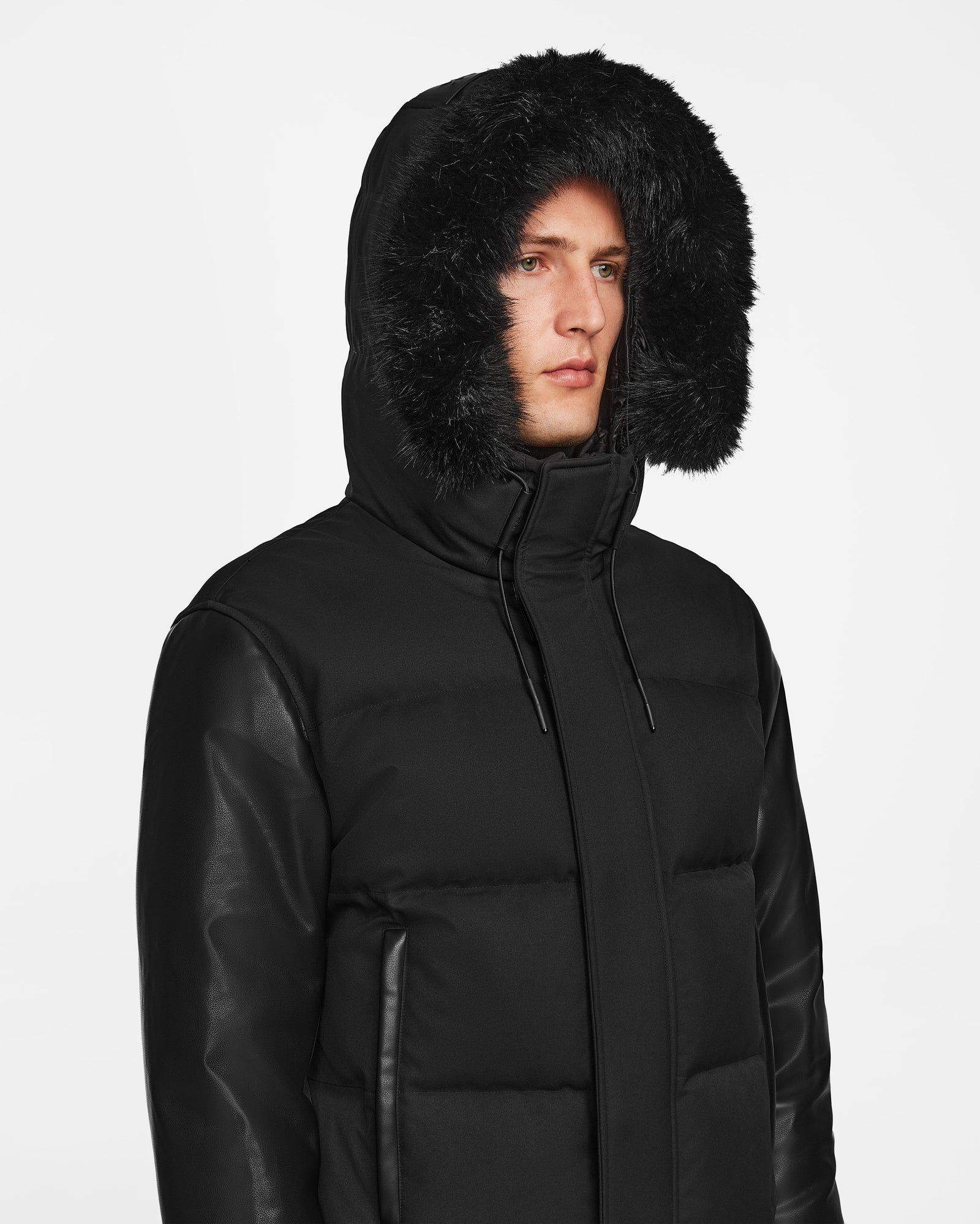 A person wearing the Rudsak DALLAS R - M123501 BLACK: a men's quilted down bomber with recycled leather sleeves and a black fur-lined hood. Its high collar and weather-resistant design stand out against a plain white background.