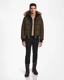 A man faces forward against a white background, wearing the Rudsak DALLAS - M122501 Army weather-resistant puffer jacket with fur-trimmed hood, plus a beige sweater, black pants, and brown lace-up boots.