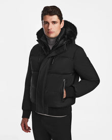 A man models the Rudsak DALLAS - M122501 Black insulated puffer jacket with a fur-lined hood, standing against a plain white background with one hand in his pocket and a neutral expression.