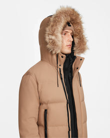 A person wears the Rudsak DALLAS - M122501 Sand, an RDS-certified tan down puffer jacket with a large fur-trimmed hood and black zipper details, looking to the side against a plain white background.