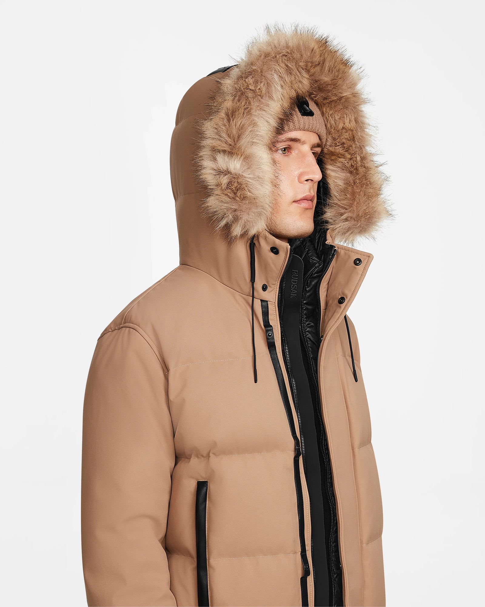 A person wears the Rudsak DALLAS - M122501 Sand, an RDS-certified tan down puffer jacket with a large fur-trimmed hood and black zipper details, looking to the side against a plain white background.