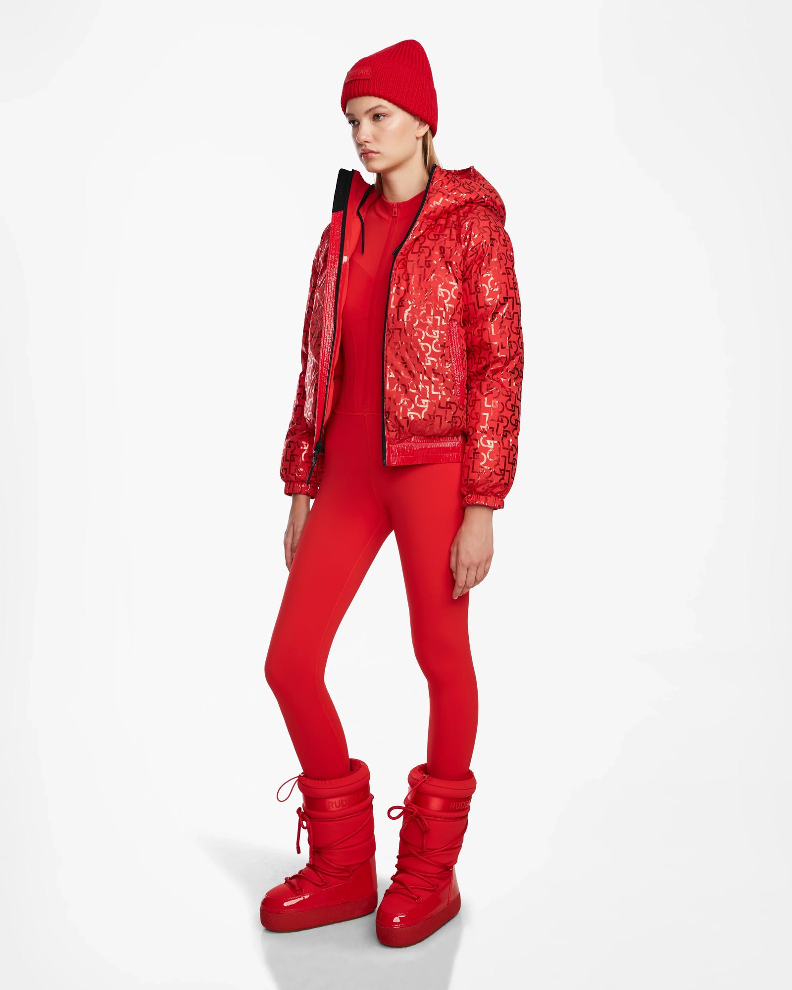A person models the Rudsak DANI M - 8124539 Flame Red, featuring a shiny red bomber-style ski jacket, beanie, bodysuit, and snow boots against a plain white background.