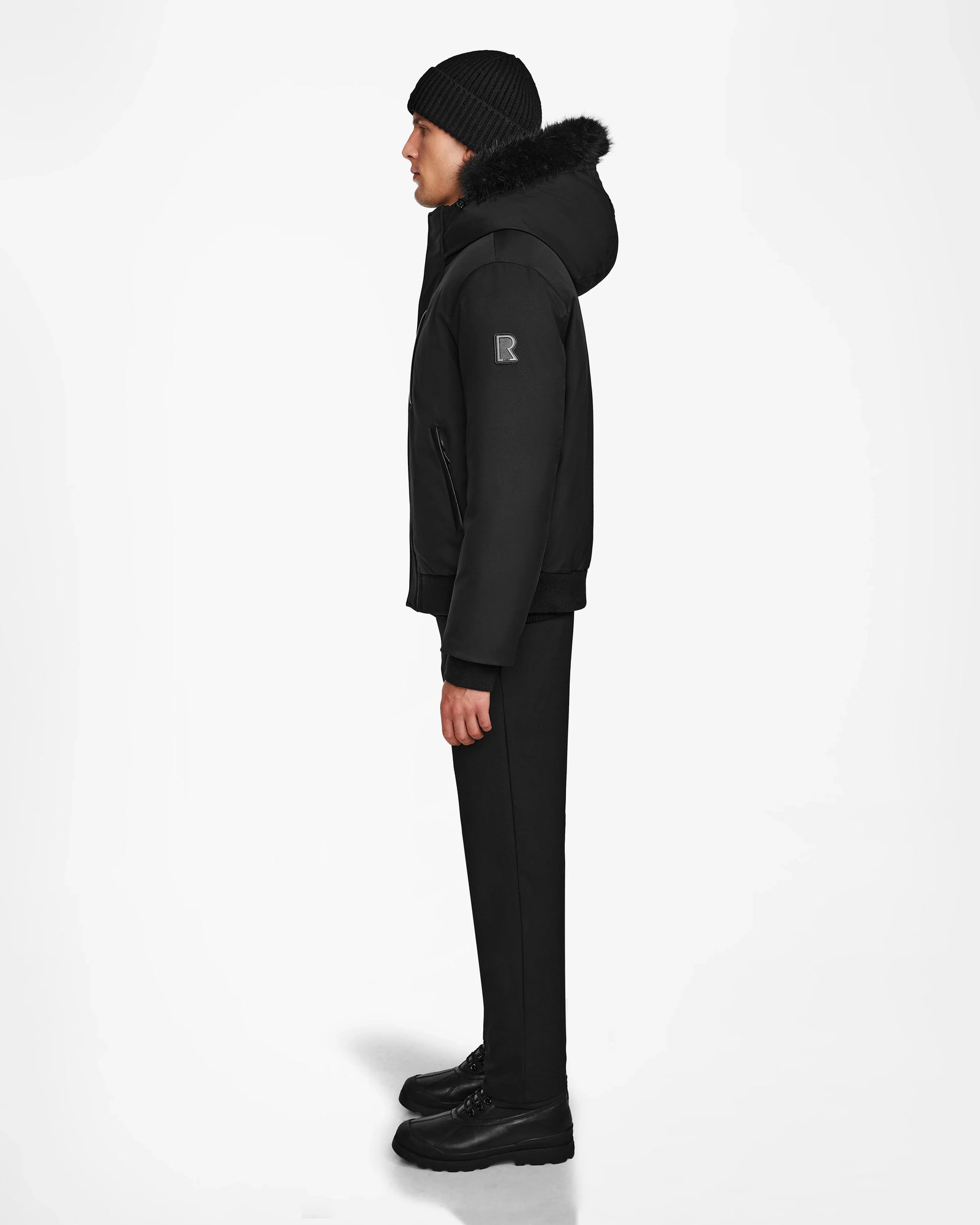 A man in profile against a white backdrop wears the Rudsak DEACON - M123513 Black/Black: a mens hooded black bomber down puffer with fur trim, paired with black beanie, pants, and shoes. The jacket is filled with RDS-certified down for warmth.