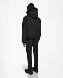 A person faces away, wearing Rudsak’s DEACON - M123513 Black/Black mens winter coat—a black hooded bomber down puffer with fur trim—paired with a black beanie, pants, and shoes against a plain white background.