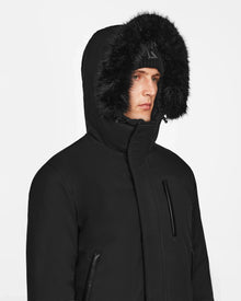 A person models the Rudsak DEACON - M123513 Black/Black bomber-style down puffer with a fur-lined hood and black beanie, gazing to the side against a plain white background.