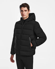 A man with short brown hair models the Rudsak DEMIAN M-R - M123504 Black men’s down puffer jacket with a hood, made from sustainable recycled fabric, standing against a plain white background, looking at the camera with one hand in his pocket.