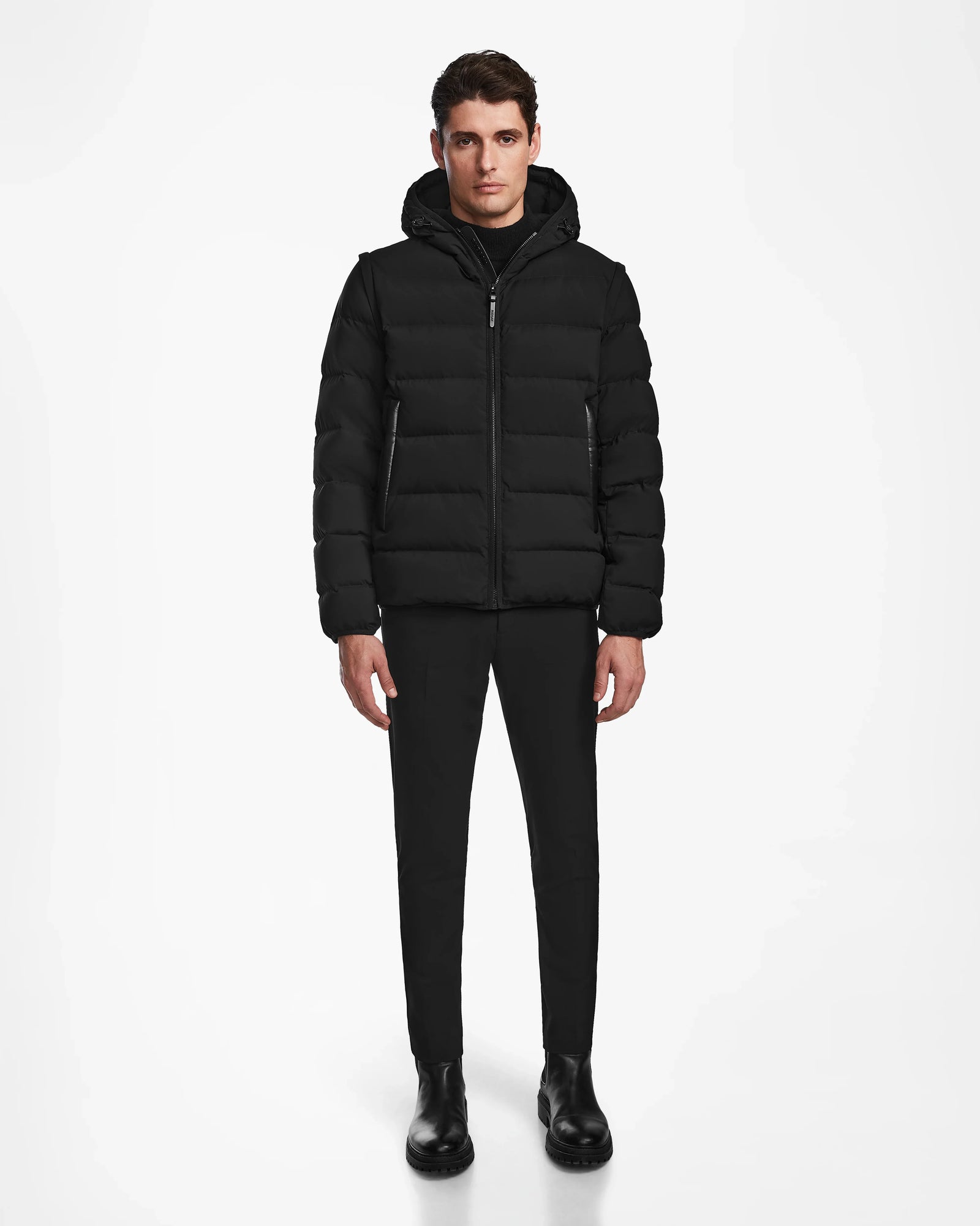 A man faces forward wearing the Rudsak DEMIAN M-R - M123504 Black mens down puffer, paired with black pants and boots, set against a plain white background.
