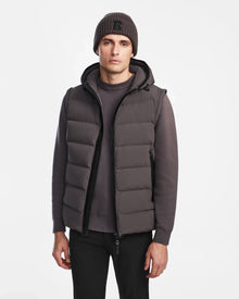 A man wears the Rudsak DEMIAN M-R - M123504 Grey down puffer vest over a gray sweater, black pants, and a knit beanie, standing against a plain white background facing forward with a neutral expression.